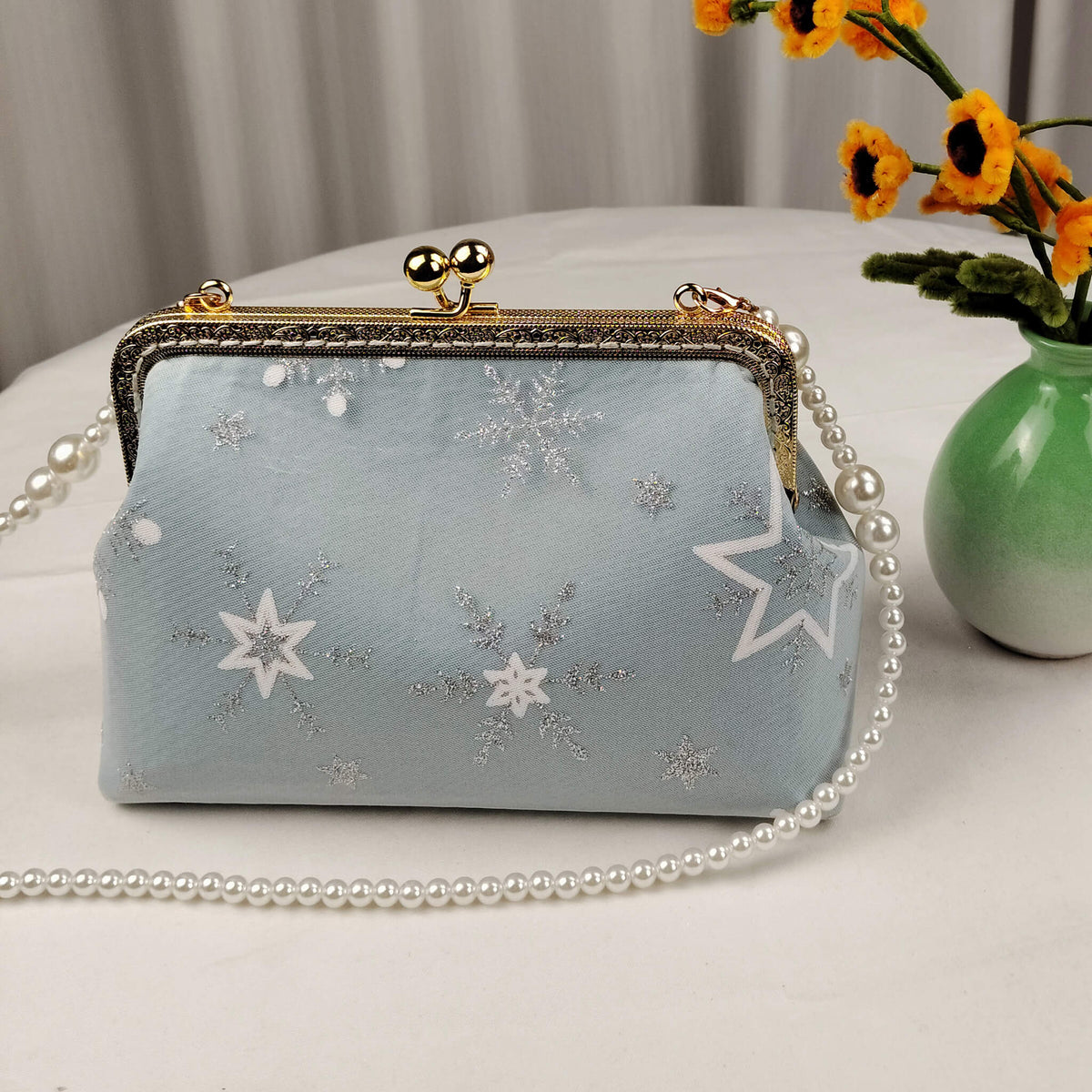Acever A002 snowflake clutch bag lifestyle shot on white table with sunflowers in green vase showing powder blue body with silver glitter snowflakes and pearl bead strap