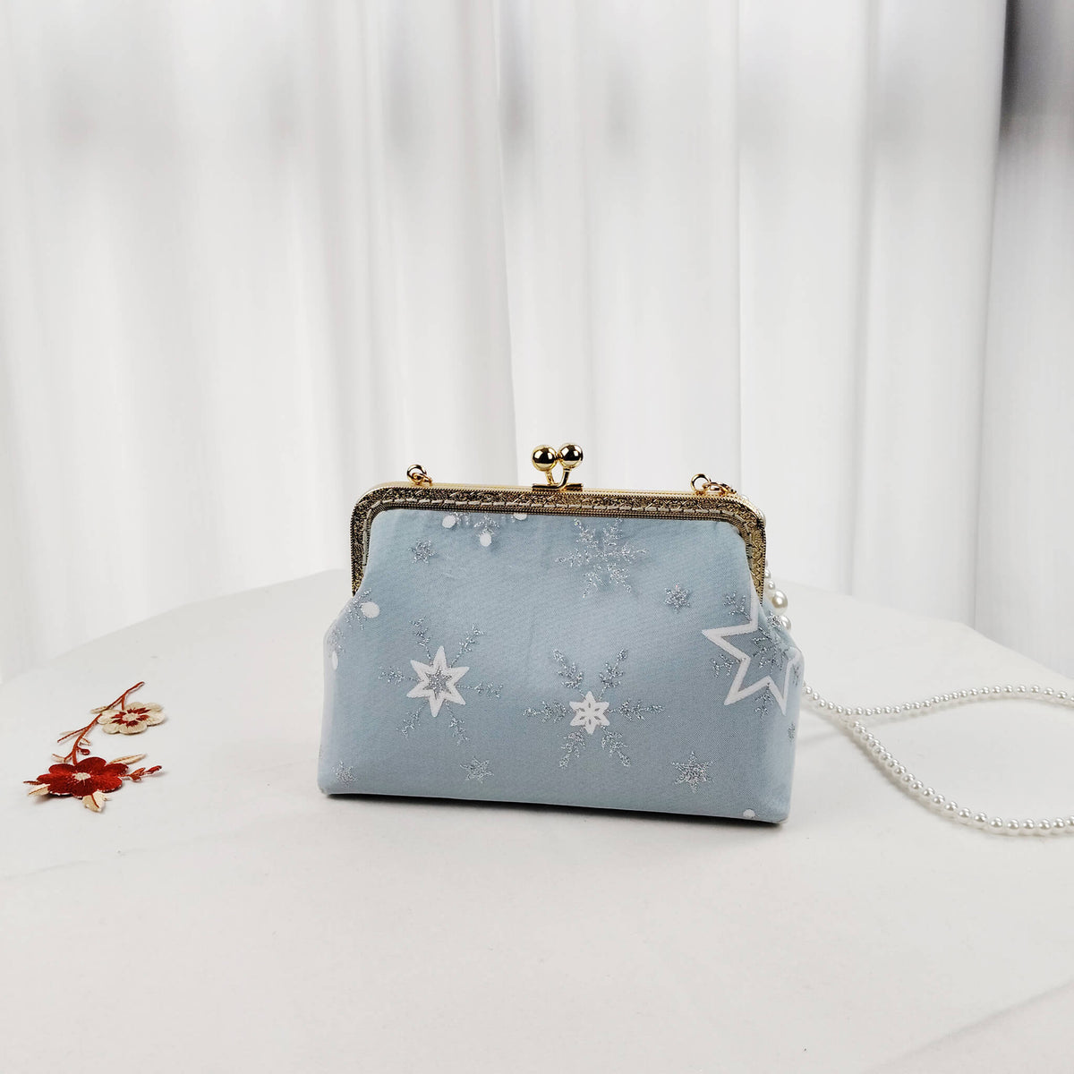 Acever A002 snowflake clutch bag lifestyle shot on white surface with red floral accessory beside showing full bag front view with gold kiss-lock frame and pearl strap