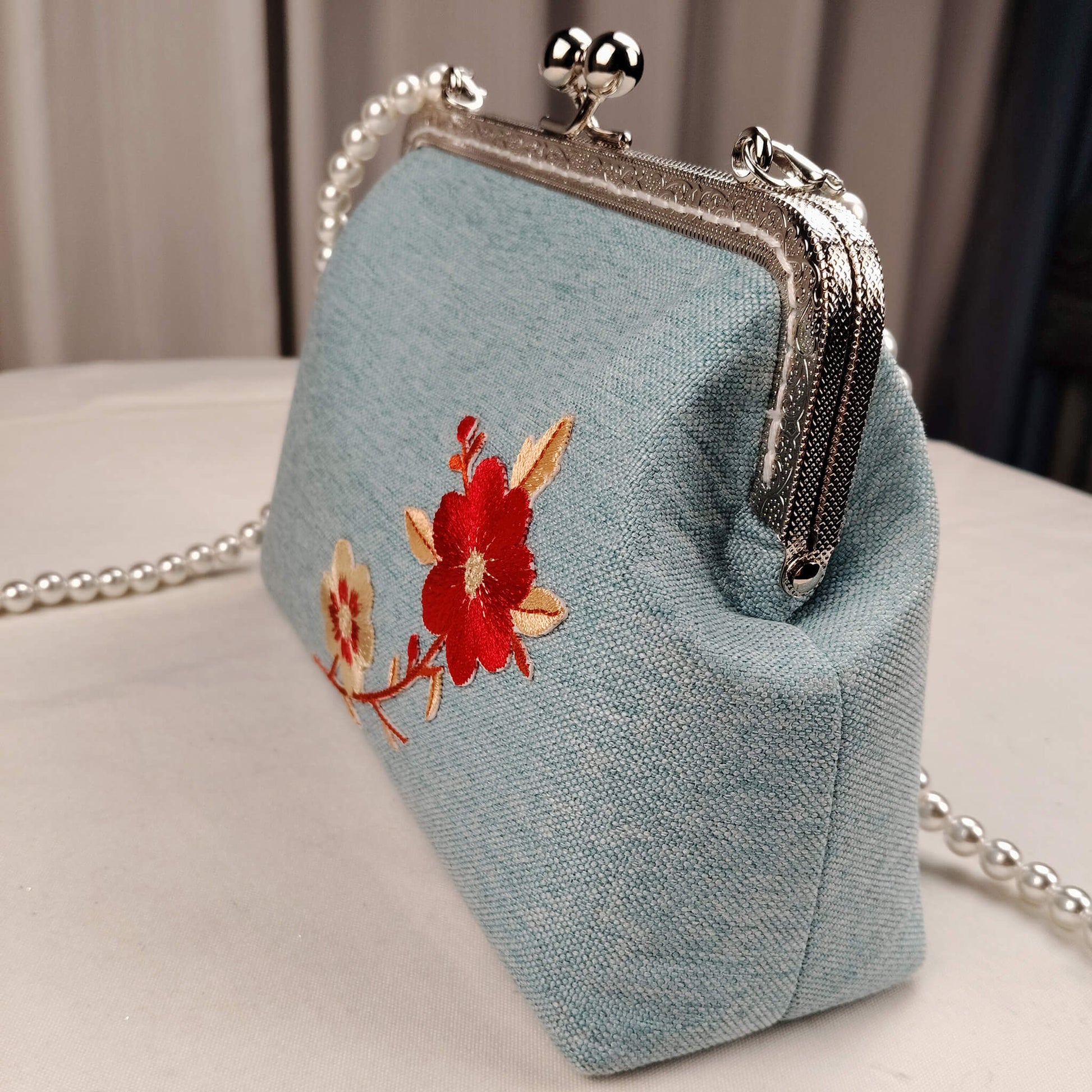 Acever A003 cherry blossom clutch bag lifestyle shot on white table showing full bag with long pearl bead crossbody strap laid out and red cream blossom embroidery on powder blue body