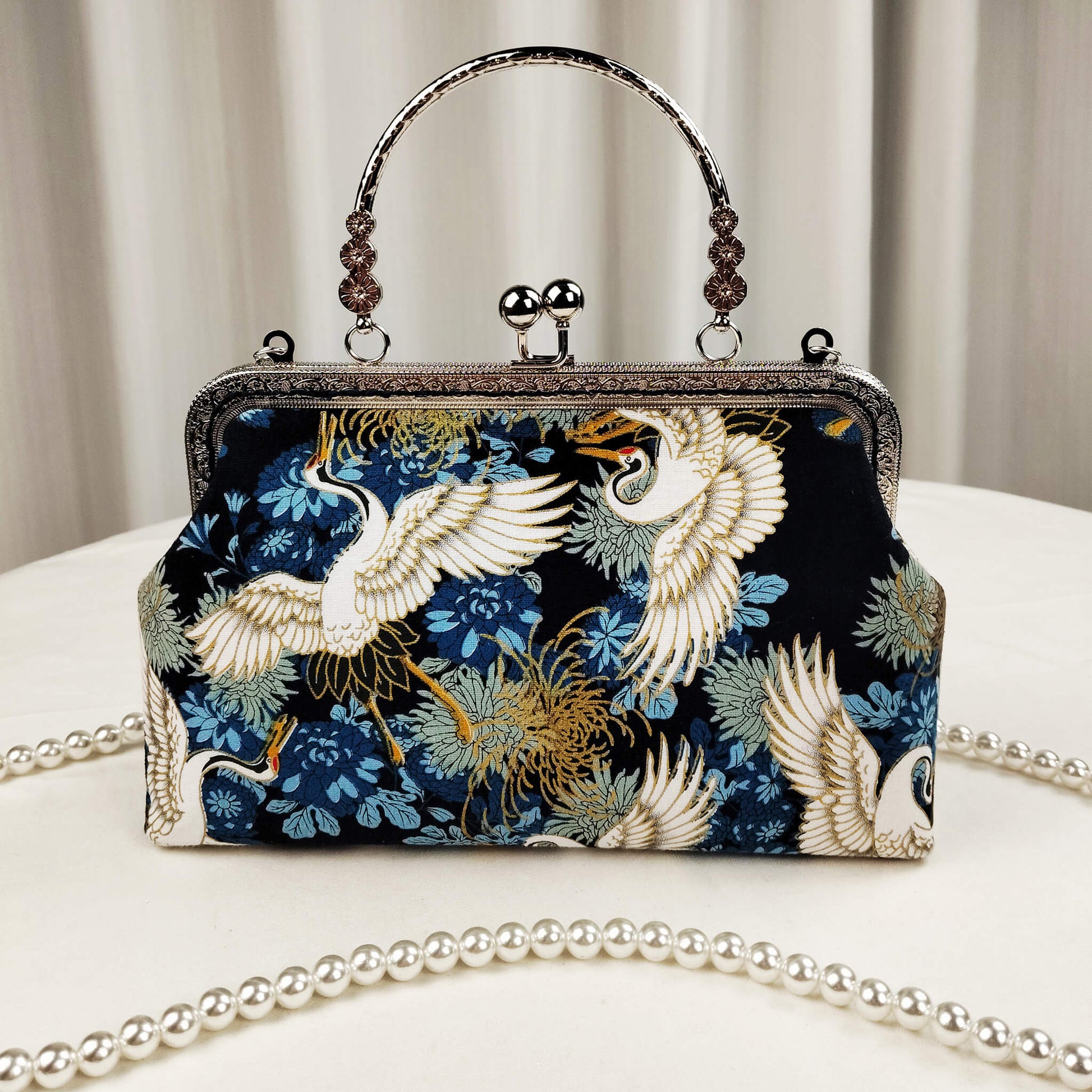Acever A006 crane print top handle bag lifestyle shot on white round table with pearl bead crossbody strap laid out showing full front view with silver kiss-lock frame and decorative top handle