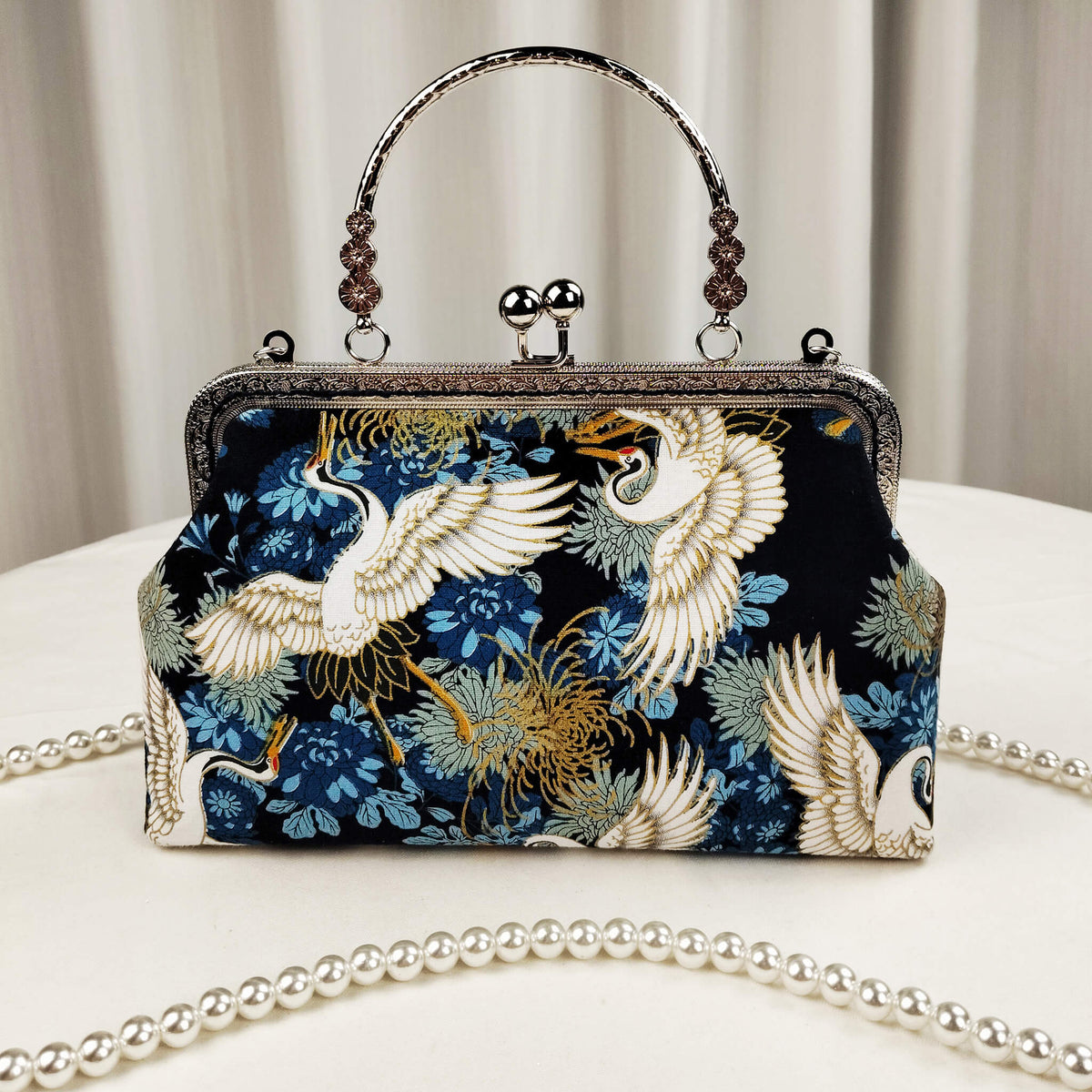 Acever A006 crane print top handle bag lifestyle shot on white round table with pearl bead crossbody strap laid out showing full front view with silver kiss-lock frame and decorative top handle