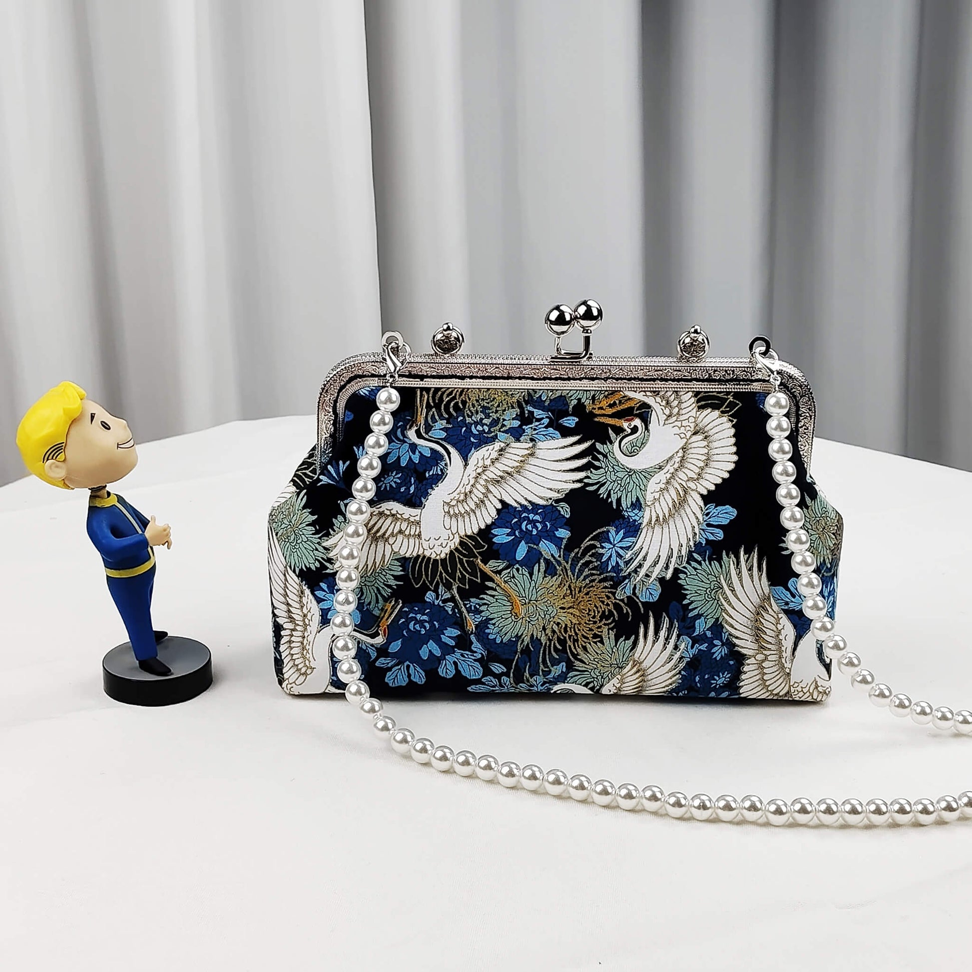 Acever A006 crane print top handle bag lifestyle shot on white table beside small yellow figurine showing front view with pearl bead strap and silver ornate kiss-lock frame