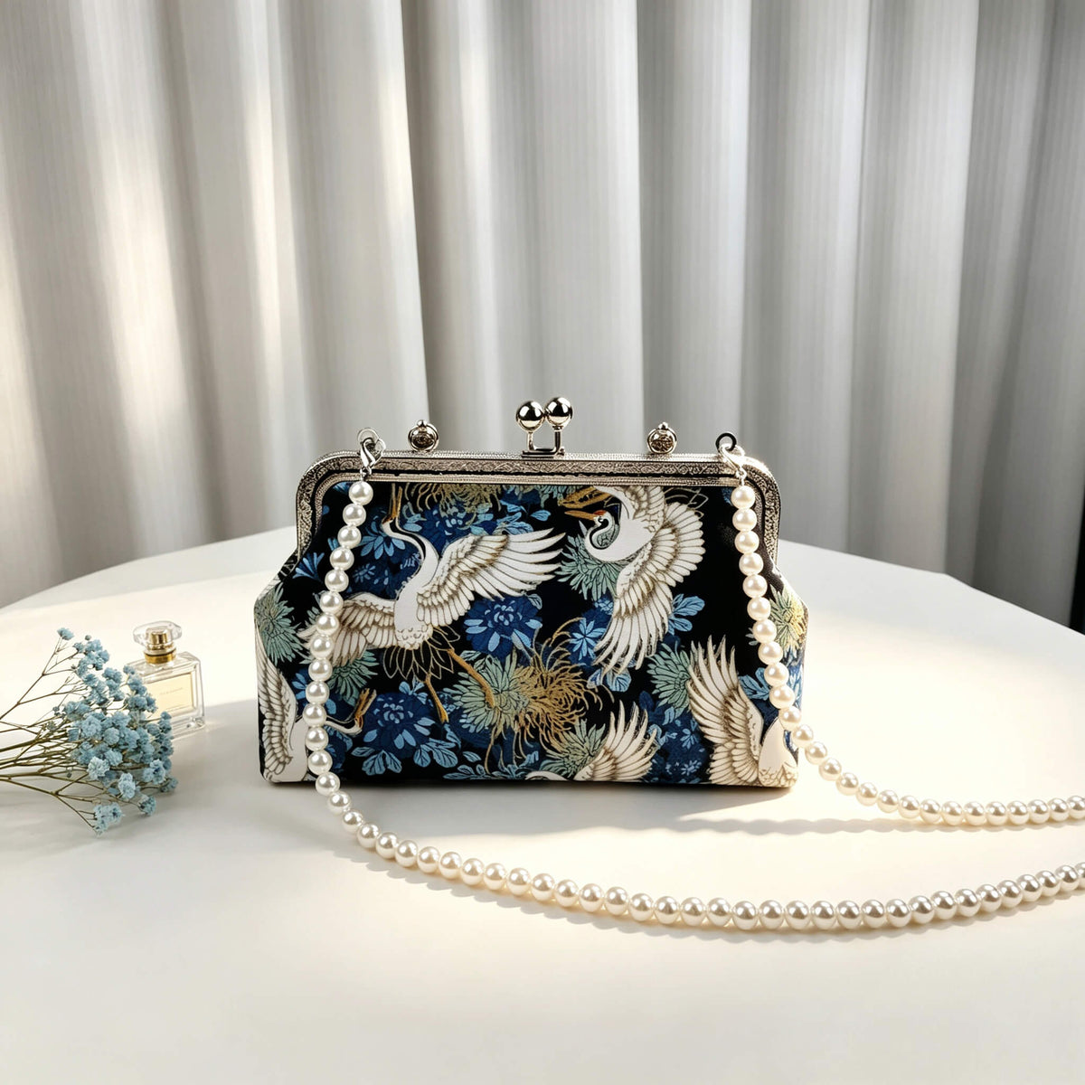 Acever A006 crane print top handle bag lifestyle shot on white table beside blue baby's breath flowers and perfume bottle showing full front view with pearl bead strap and silver top handle