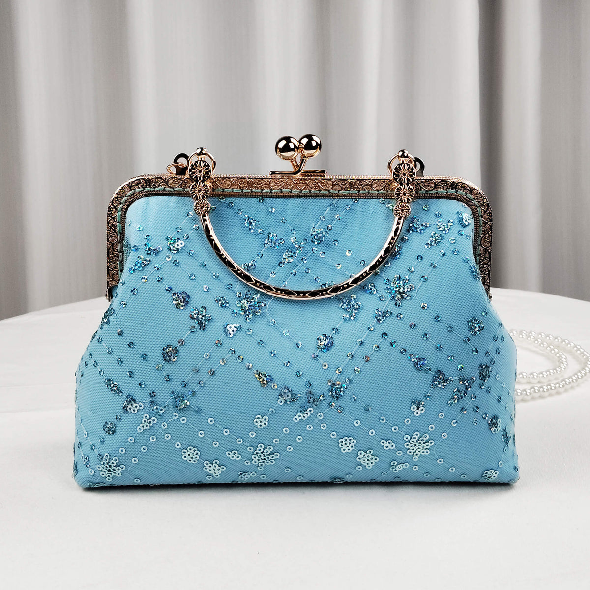 Acever A007 blue sequin floral clutch bag lifestyle shot on white round table against grey curtain backdrop showing full front view with antique gold kiss-lock frame curved top handle and pearl bead crossbody strap