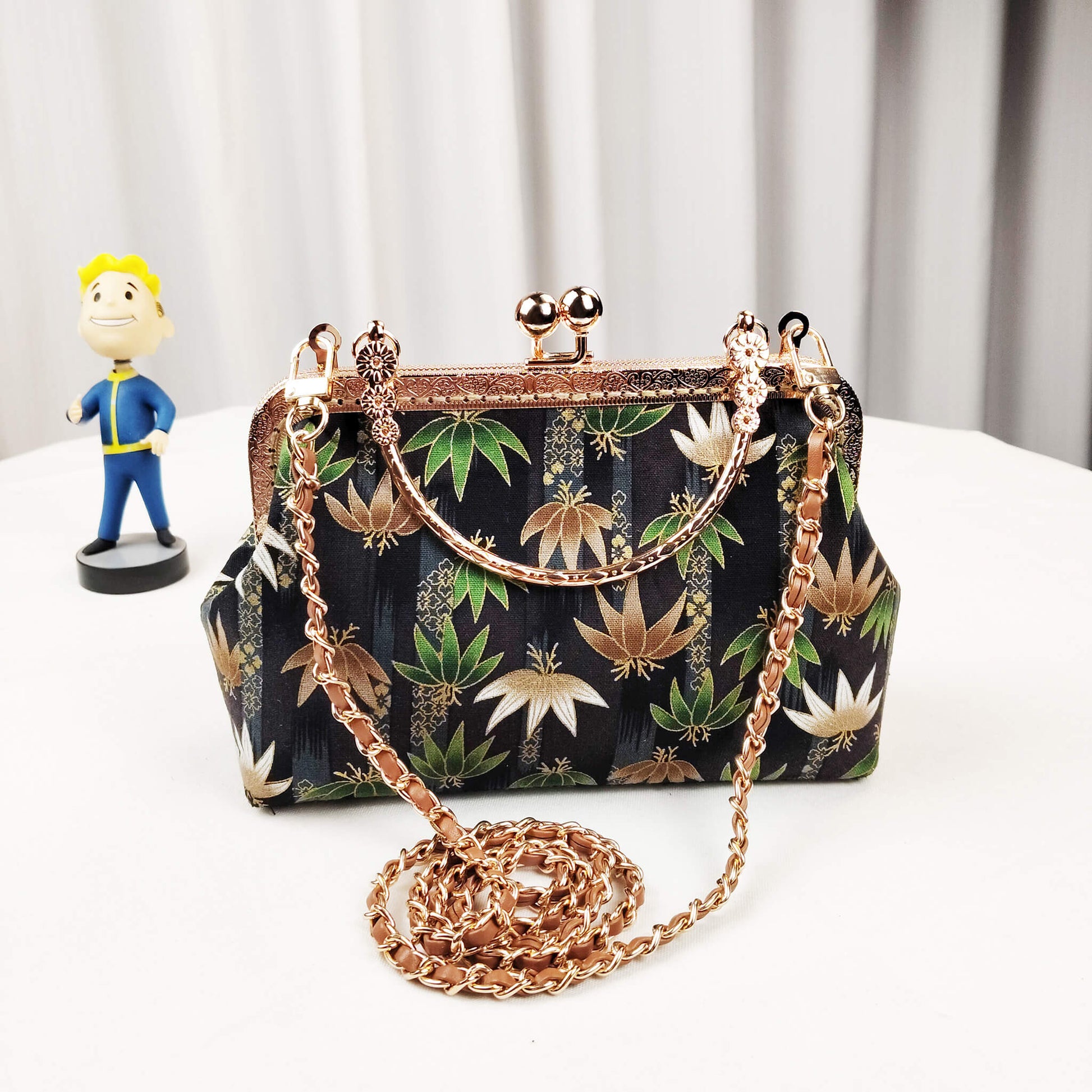Acever B002 lotus and leaf jacquard top handle bag lifestyle shot on white round table beside Fallout Vault Boy figurine showing full front view with rose gold kiss-lock frame decorative top handle and long rose gold chain crossbody strap