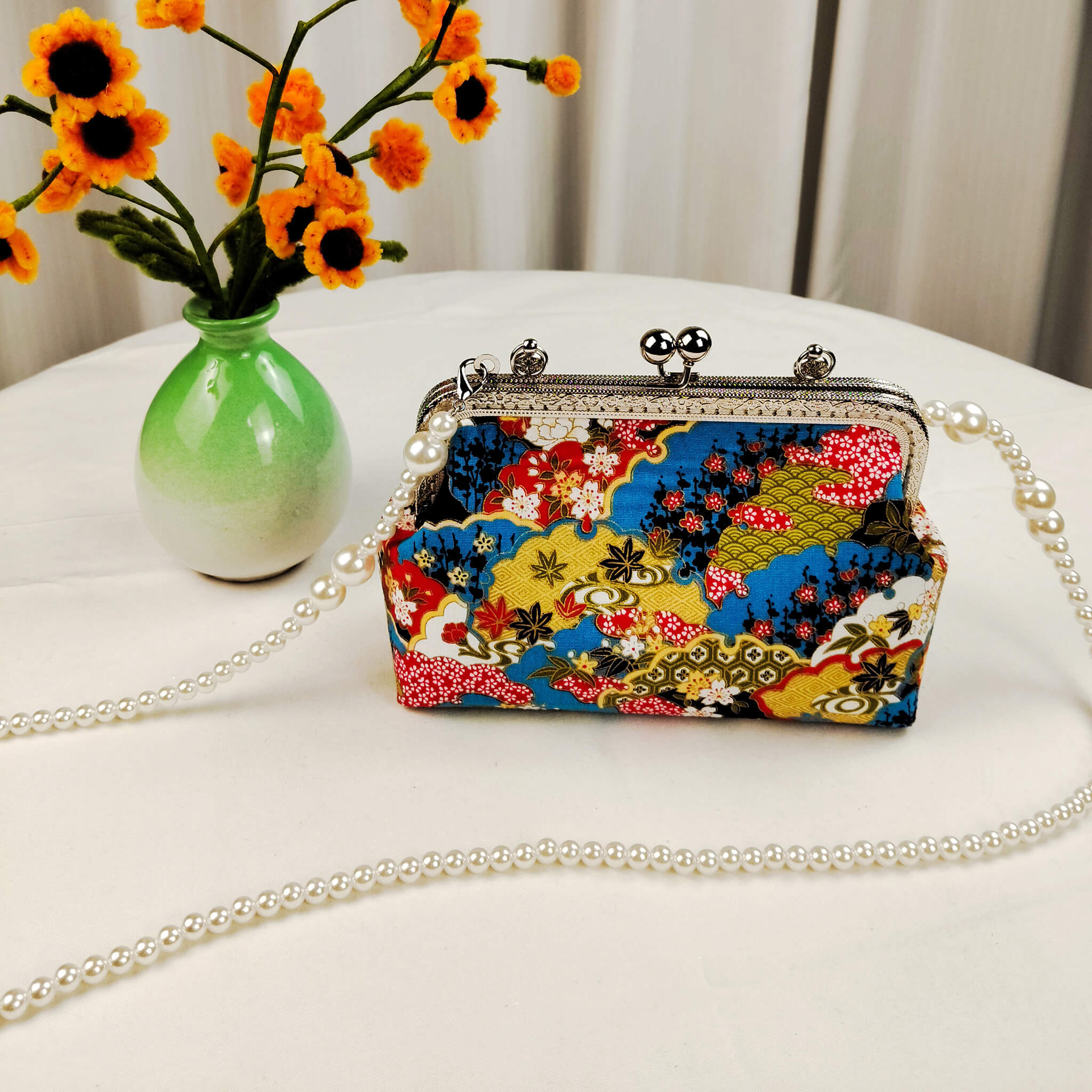 Acever B003 Japanese Nishiki brocade top handle bag lifestyle shot on white round table beside green ceramic vase with orange sunflowers showing front view with antique silver kiss-lock frame and large pearl bead crossbody strap