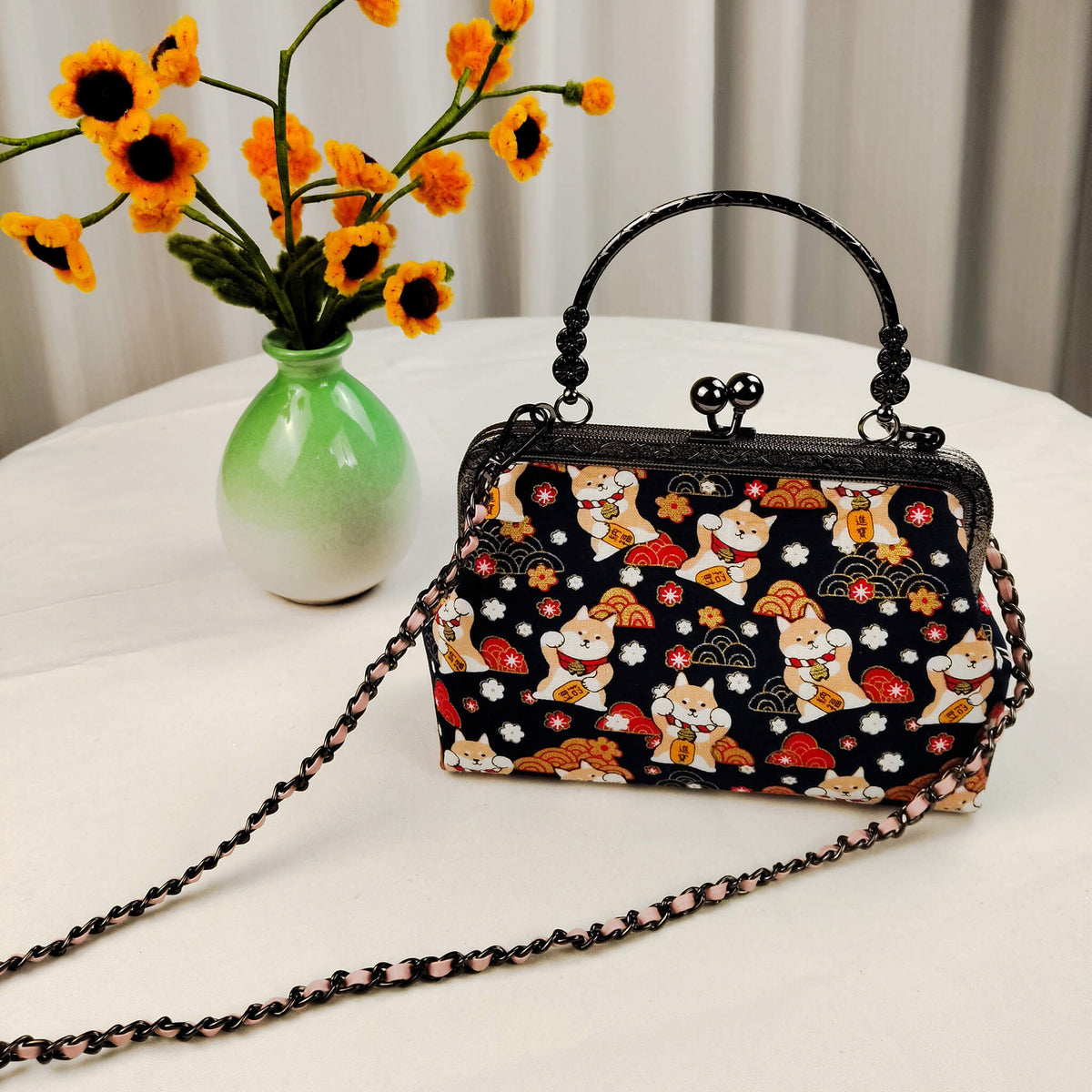 Acever B005 Lucky Shiba Inu top handle bag lifestyle shot on white round table beside green ceramic vase with orange sunflowers showing full front view with gunmetal kiss-lock frame crystal bead top handle and blush pink woven gunmetal chain crossbody strap