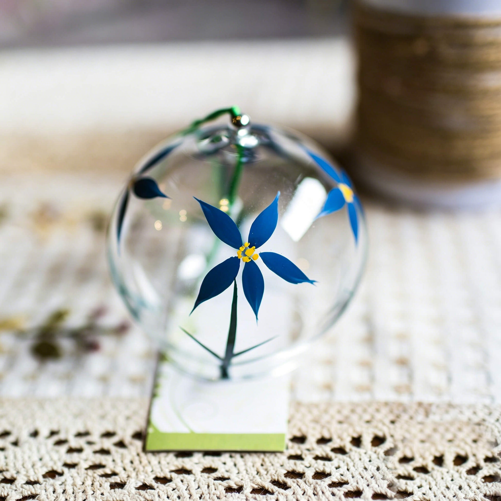 Close-up of Acever blue daisy glass wind chime X23 hanging against stone wall showing clear globe with blue daisy blossoms, yellow centres, green stems, and white vine paper strip