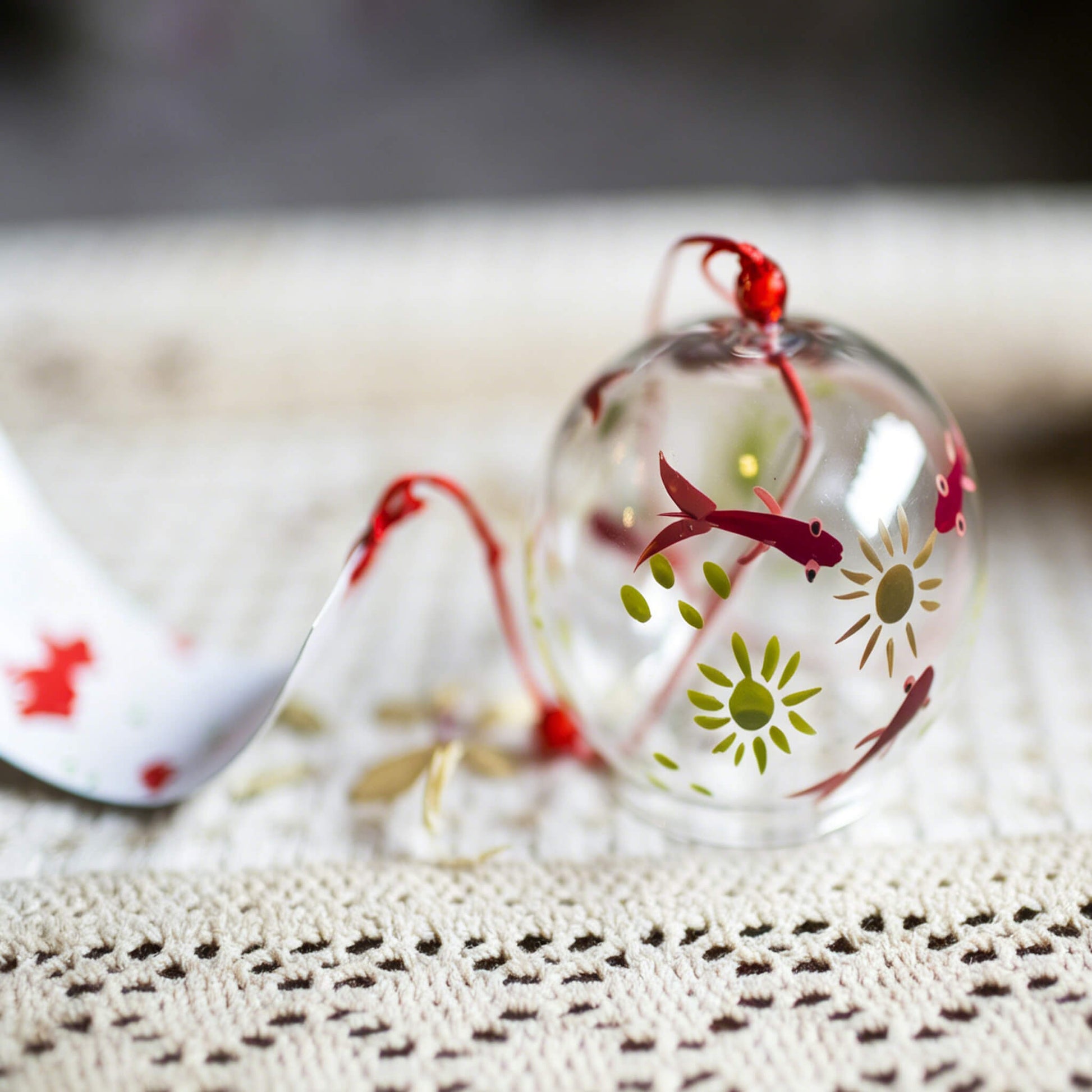 Close-up of Acever red goldfish glass wind chime X27 nested on knitted mat  showing clear globe with red kingyo fish and green chrysanthemum bloom