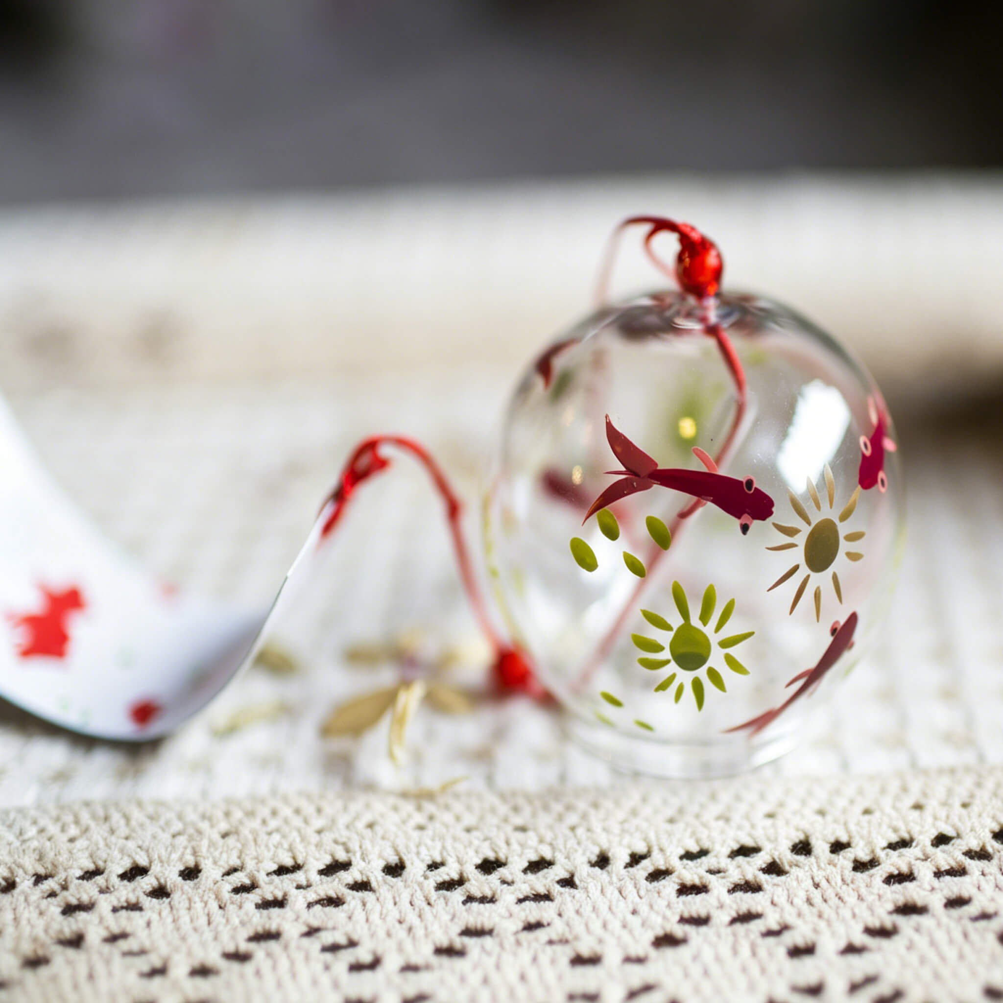 Close-up of Acever red goldfish glass wind chime X27 nested on knitted mat  showing clear globe with red kingyo fish and green chrysanthemum bloom