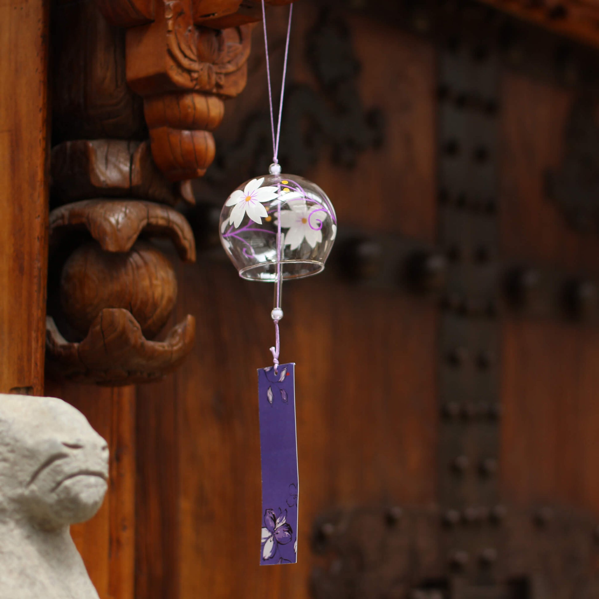 Acever Japanese furin glass wind chime X28 hanging on traditional Chinese temple carved wooden piilar and stone ram sculpture showing clear globe with white magnolia blossoms and magenta vine scrolls with deep violet iris paper strip. 
