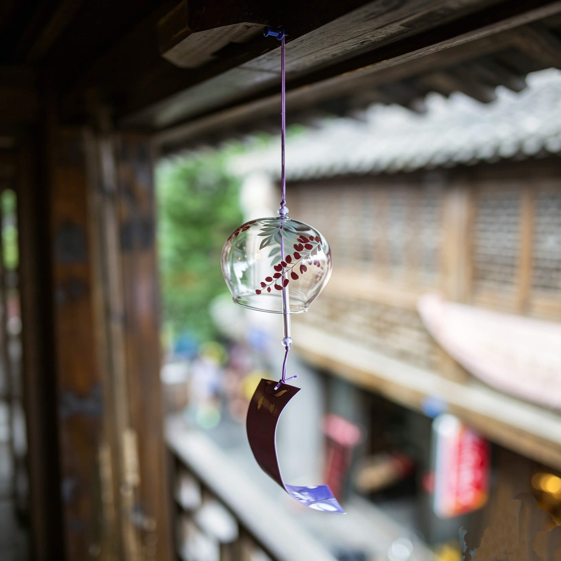 Acever purple wisteria glass wind chime X30 hanging from traditional Japanese wooden eave in old street showing clear globe with green fern fronds and burgundy wisteria petals and dark curled paper strip