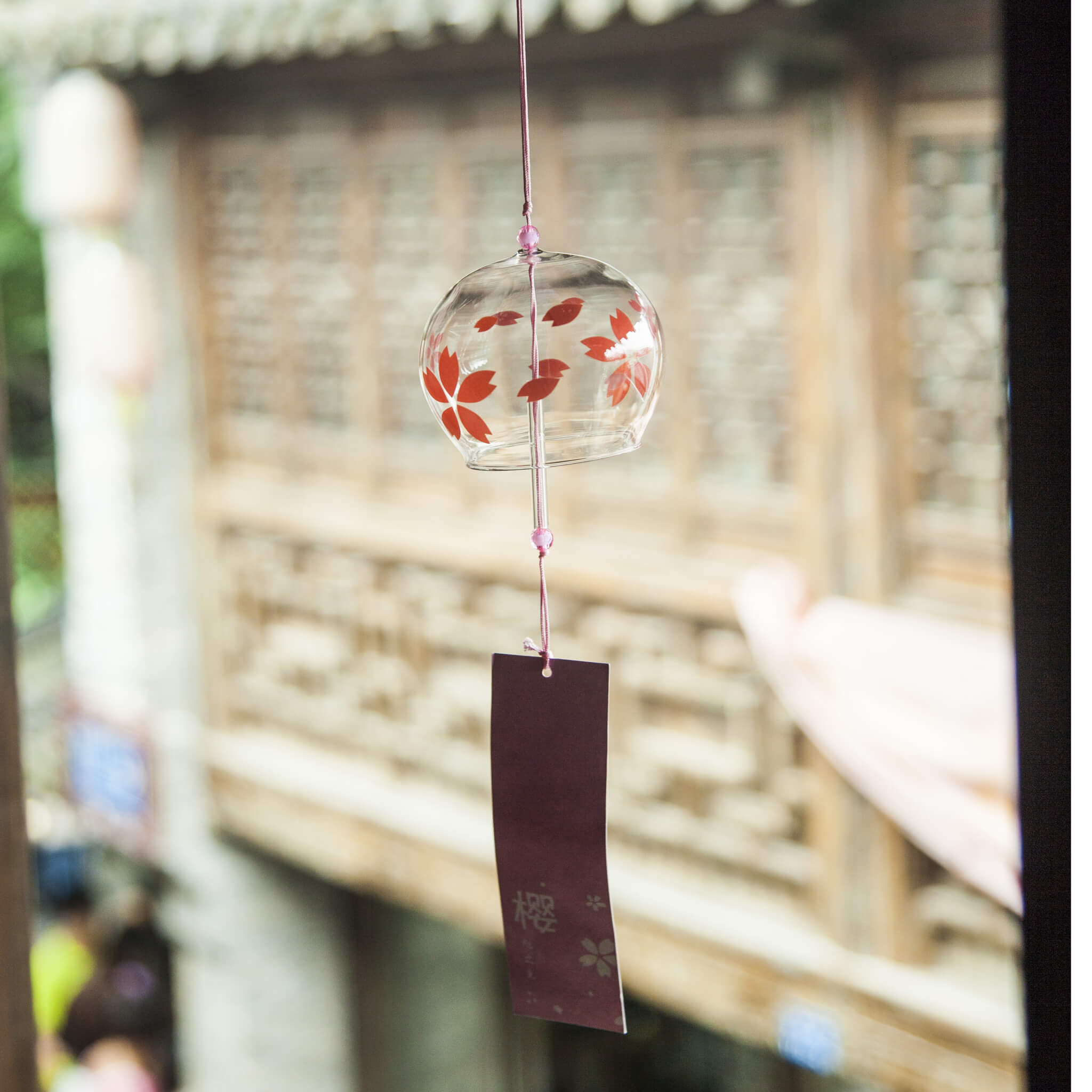 Acever red sakura glass wind chime X37 hanging from traditional Japanese wooden eave in old street showing clear globe with red sakura blooms and dark burgundy paper strip with sakura kanji