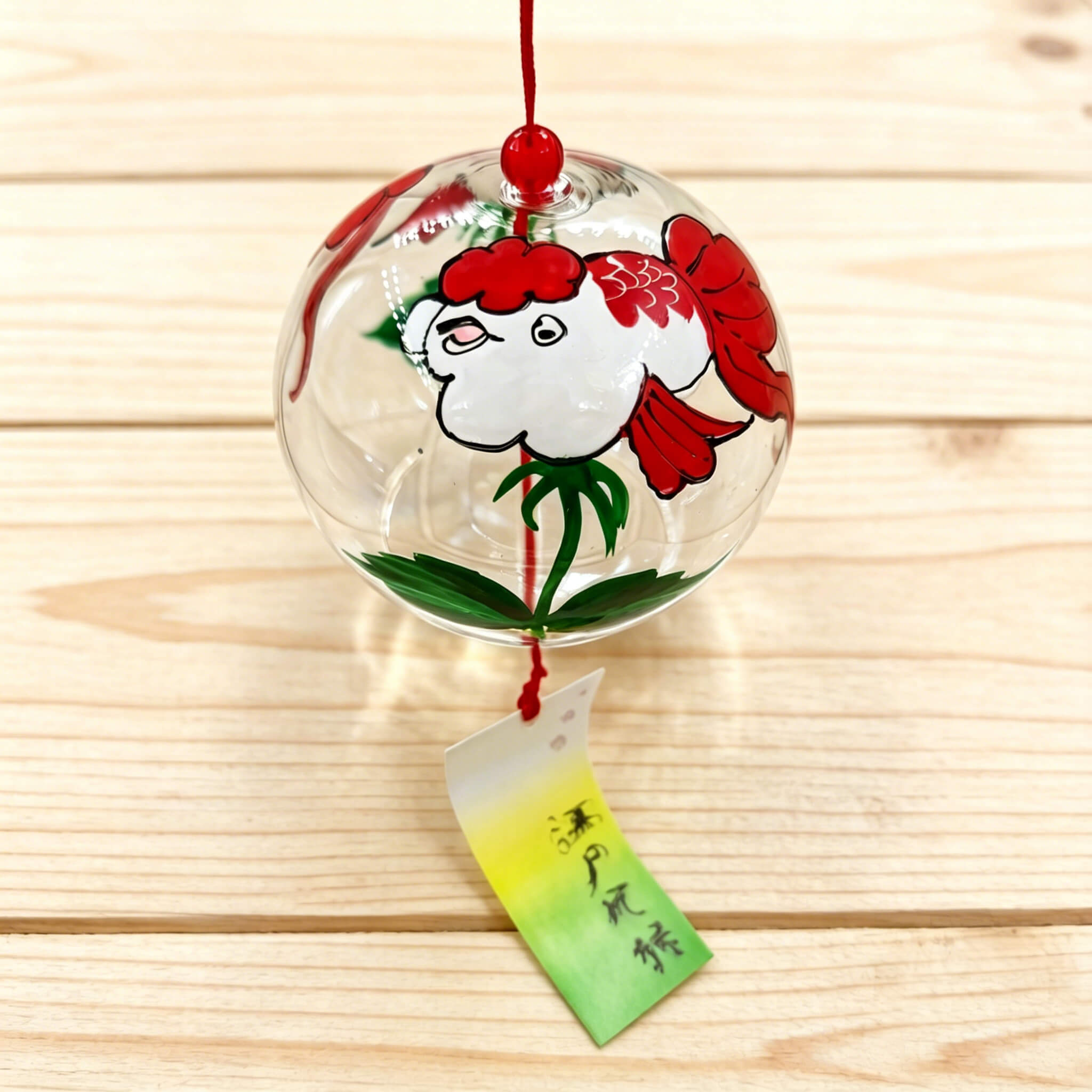 Right view of Acever goldfish glass wind chime X39 on wooden plank surface showing clear ribbed globe with red and white kingyo goldfish, green aquatic leaves, and white-to-green Edo Minyo paper strip