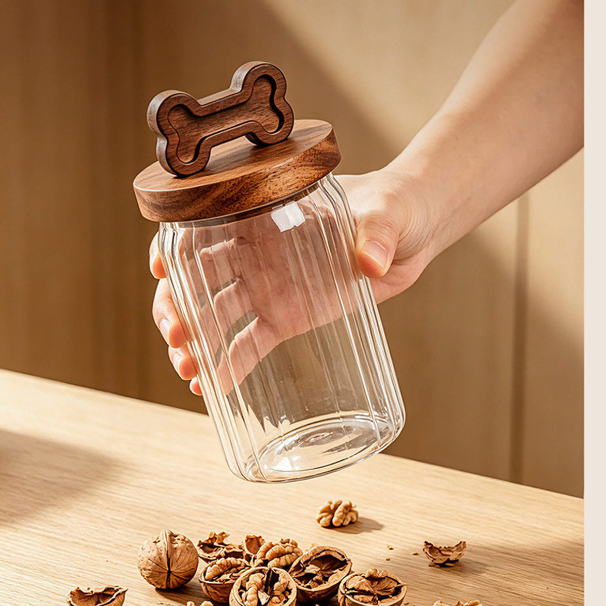 Hand holding ribbed glass jar with bone-handle wooden lid above cracked walnuts.