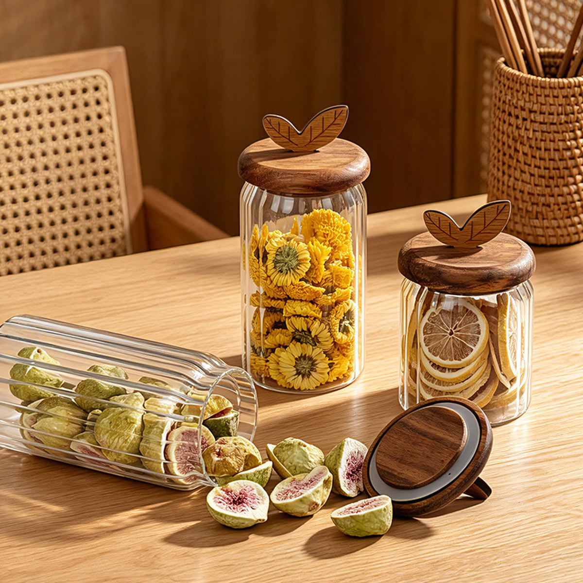 Three ribbed glass jars with leaf lids on wooden shelf filled with dried figs, flower tea, and dried lemon slices.
