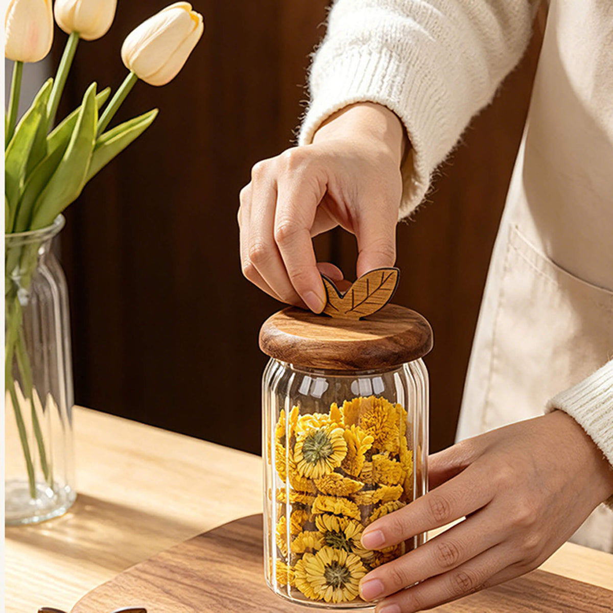 Three ribbed glass jars with leaf lids on woven round placemat filled with dried figs, flower tea, and lemon slices.